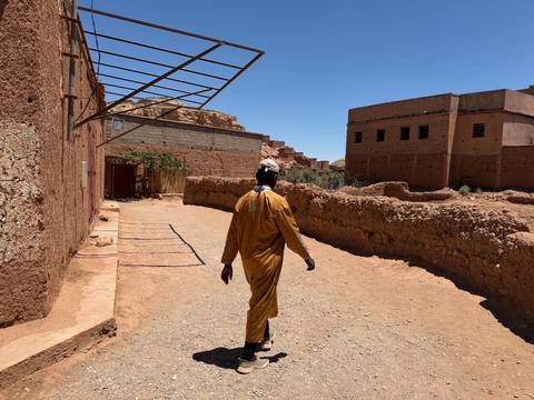       Man in traditional robe walking a dusty lane lined with earthen walls in a desert village.
  