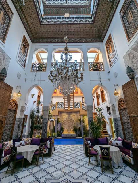       Opulent Moroccan riad courtyard with intricate mosaics, carved wood and crystal chandelier.
  