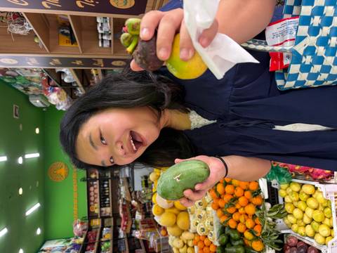       Smiling woman holds tropical fruits while browsing a colorful Vietnamese grocery shop.
  