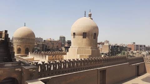       Twin domes and crenellated walls of an old Cairo mosque overlook densely packed city buildings.
  