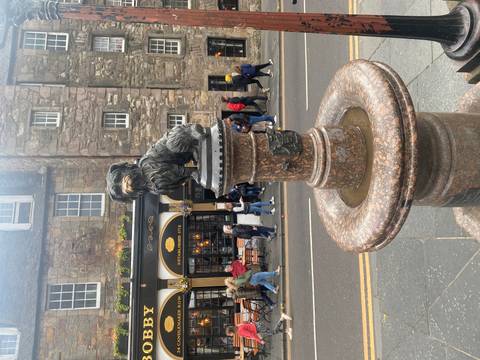       Bronze statue of Greyfriars Bobby on pedestal with busy Edinburgh street behind.
  