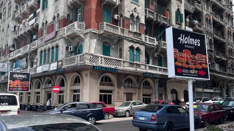       Busy city corner with historic building featuring green shutters, billboard advertising burgers and several parked cars.
  