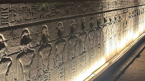       Dimly lit corridor of hieroglyphic wall reliefs illuminated by warm accent lighting.
  