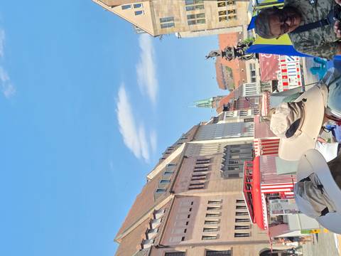       Busy outdoor scene with tourists in sun hats looking toward historic buildings and a church spire.
  