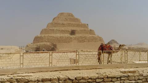       Step Pyramid of Djoser at Saqqara with a saddled camel standing beside a stone fence in the desert haze.
  