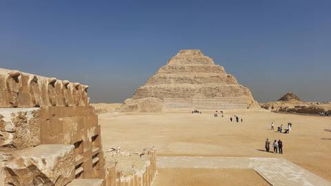       Wide open courtyard view of the Step Pyramid of Djoser with small groups of tourists scattered across the sand.
  