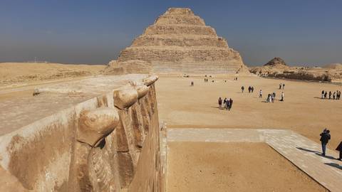       Wide desert courtyard leading to the Step Pyramid of Djoser with small tourist groups scattered around.
  