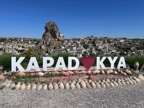       Large 'KAPADOKYA' sign with heart symbol frames Uchisar Castle and the town behind it under clear blue sky.
  