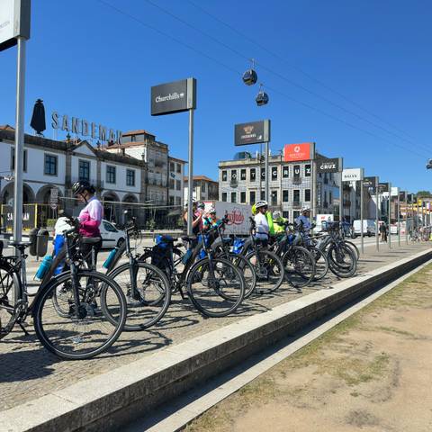       Cyclists line up with their bikes near riverfront wine warehouses and signage in Porto.
  