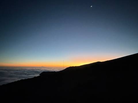       Silhouetted mountain ridge above a sea of clouds with a colorful sunrise glow on the horizon.
  