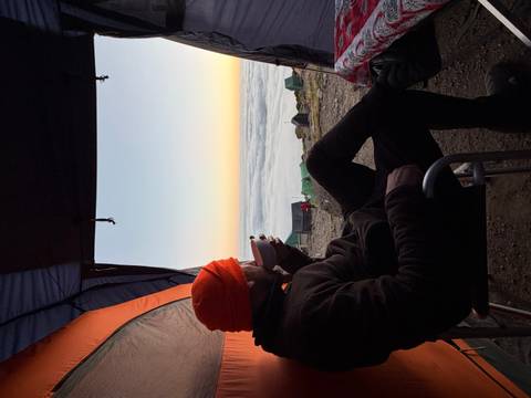       Camper relaxing with a hot drink inside a tent overlooking a cloud-filled valley at dawn.
  