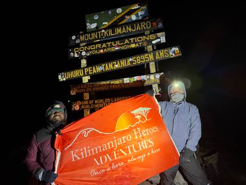       Two climbers posing with a banner at the famous Uhuru Peak sign on Mount Kilimanjaro under night skies.
  