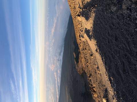       Rocky volcanic slope leading down to clouds with vast horizon and blue sky.
  