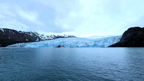       Blue tidewater glacier spills into the sea between dark cliffs and snowy peaks.
  