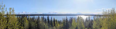       Sparkling lake bordered by conifer forest with distant snow-capped range under bright sky.
  