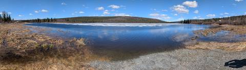      Blue lake partly covered by melting ice surrounded by low forested hills and fluffy clouds.
  