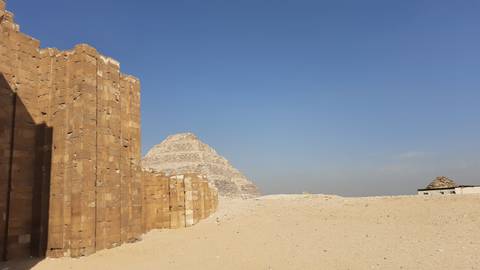       Step pyramid rising behind weathered sandstone walls amid a vast desert plateau under a clear blue sky.
  