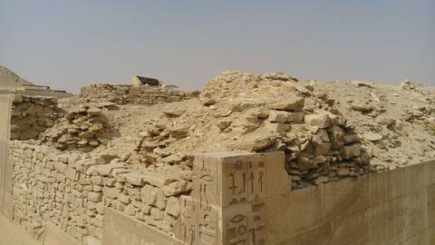       Close-up view of crumbling stone and mud-brick ruins adorned with faint hieroglyphics under hazy desert light.
  