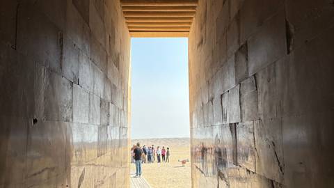       Long narrow stone corridor opening onto a bright desert plain where a tour group gathers at the exit.
  