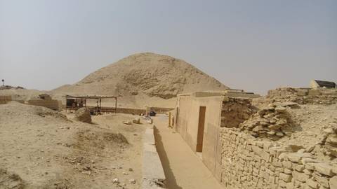       Low desert mound and ancient stone walls stretching along a sandy path beneath a pale sky.
  