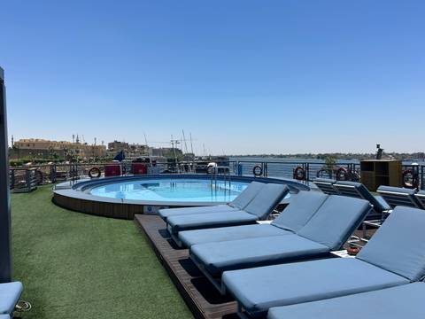       A rooftop pool with blue loungers overlooks the Nile River and distant temple ruins.
  