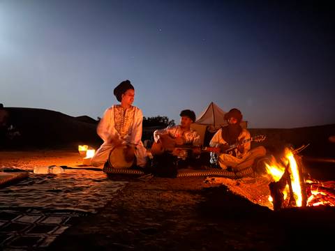       Musicians play drums and guitar beside a campfire with a night sky overhead at a desert camp.
  
