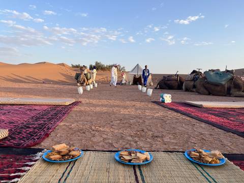       Low tables and patterned rugs await breakfast with camels and dunes in the background.
  