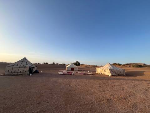       A wide desert campsite with three canvas tents sits amid rolling dunes under a clear sky.
  