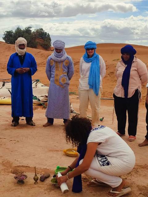       Travelers listen to a guide in traditional dress beside camel gear on desert sand.
  