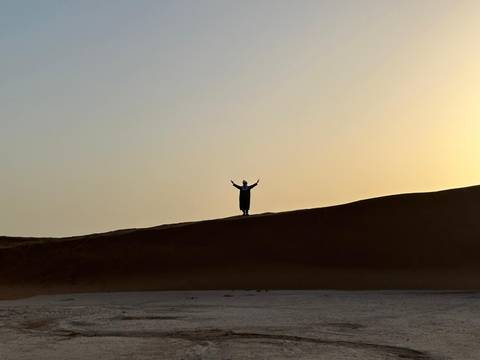       A lone silhouetted figure raises arms atop a high sand dune against a hazy desert sunset.
  