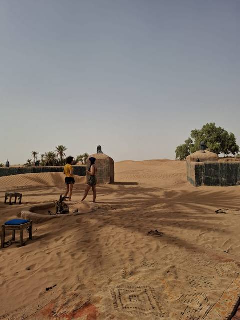       Two travellers exploring a rustic desert settlement surrounded by sand and palms.
  