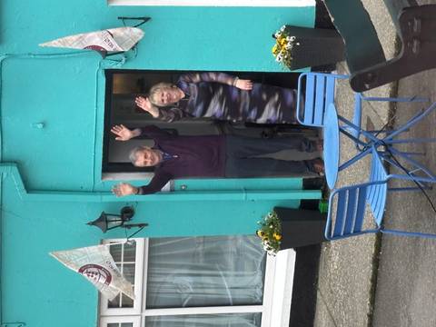       An older couple wave cheerfully from the doorway of a brightly painted turquoise house.
  