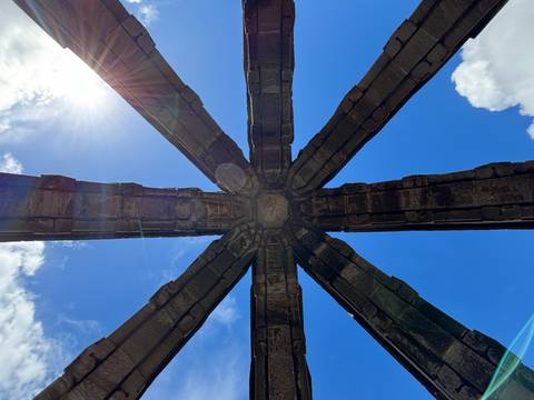       Looking straight up through intersecting stone arches of a ruined structure against a bright blue sky with sun flare.
  