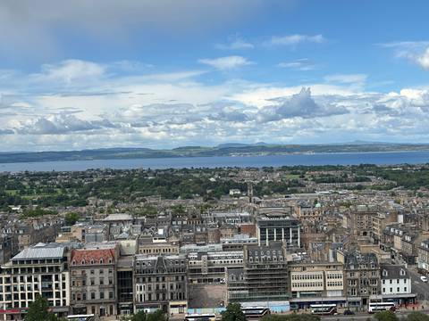       Panoramic cityscape of Edinburgh with rooftops, greenery and the Firth of Forth under patchy clouds.
  