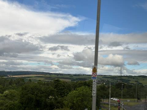       Countryside view with patchwork fields seen through a bus window, partly obscured by a pole.
  