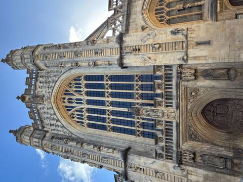       Gothic façade of Bath Abbey with towering windows, carvings and a bright blue sky background.
  