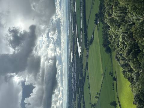       Sweeping aerial view of farmland, river bends and distant horizon beneath dramatic sun-lit clouds.
  