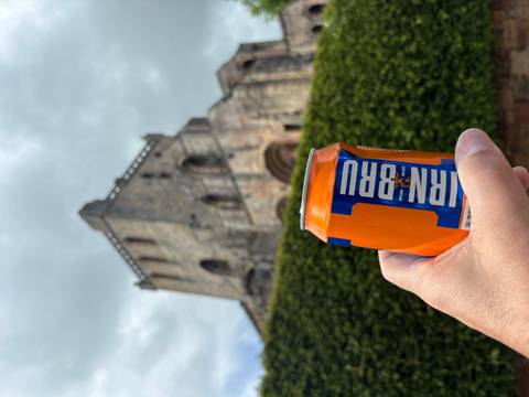       Hand holding an IRN-BRU can with a stone abbey in soft focus background.
  