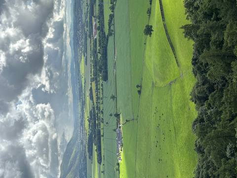       Verdant patchwork of flat fields and hedgerows stretching toward distant hills under broken clouds.
  