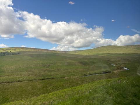      Expansive open moorland with rolling grassy hills under a bright blue sky viewed through a coach window.
  