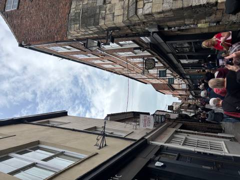       Narrow historic shopping lane crowded with visitors beneath patchy sky.
  