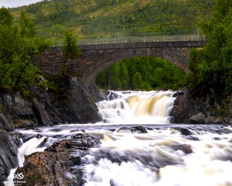       Silky waterfall plunges beneath a stone arch bridge amid lush Norwegian forest.
  