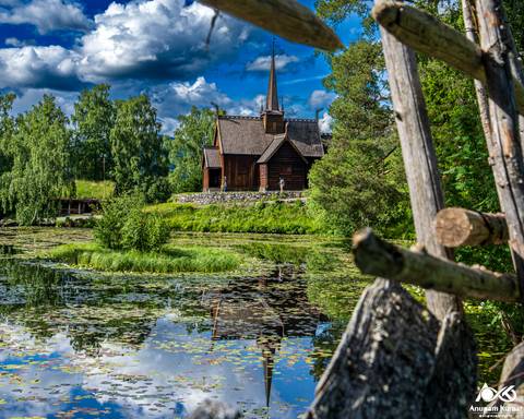       Traditional stave church reflected in a lily-covered pond surrounded by lush greenery and dramatic clouds.
  