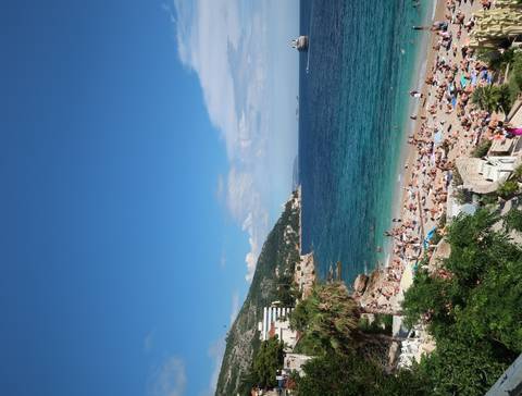       Busy Mediterranean beach packed with sunbathers and turquoise waves rolling onto golden sand.
  