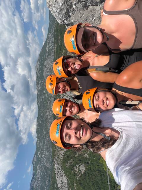       Smiling adventurers wearing orange helmets take a selfie with rugged cliffs behind.
  