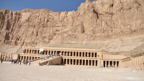       The terraced Temple of Hatshepsut nestled below tall desert cliffs with visitors walking across the forecourt.
  