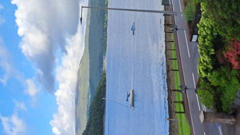       Vertical phone shot of a calm lake with sailboats and green hills under patchy clouds.
  