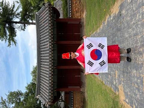       Man dressed in traditional red hanbok holding the South Korean flag in a historic courtyard.
  