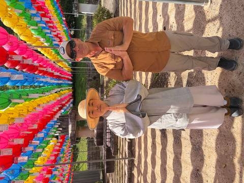       Monk and traveler standing with hands together beneath colorful lantern canopy at a temple festival.
  