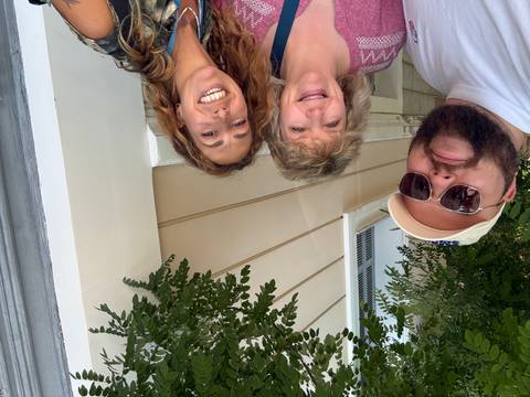       Three people smiling beneath leafy branches beside a cream-colored building
  